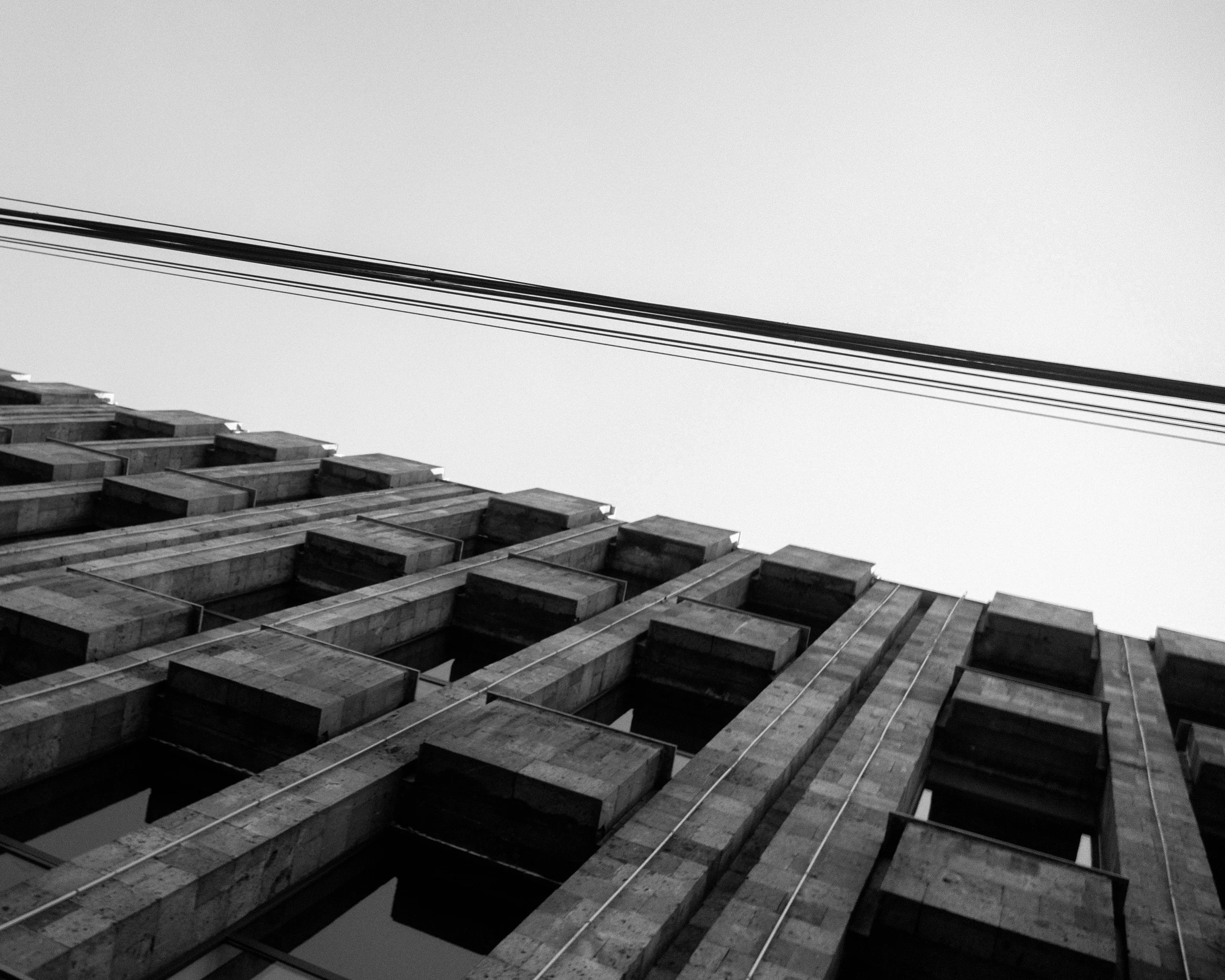 An upward view of a geometric building facade crossed by overhead wires